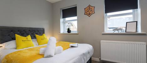 A neatly made bed with a grey headboard, white and yellow bedding, and a white towel. There are two windows with black blinds, one with a picture frame.