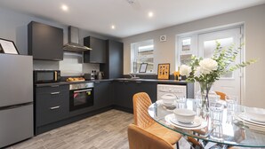 A modern kitchen with dark cabinetry, a glass dining table, and a vase with flowers. - Champagne House - 3 Bedroom Townhouse (Darlington)