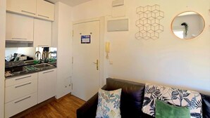 Ein gemütlicher Wohnraum mit einer Küchenzeile, einer Couch mit gemusterten Kissen, einem runden Spiegel und geometrischer Wandgestaltung.