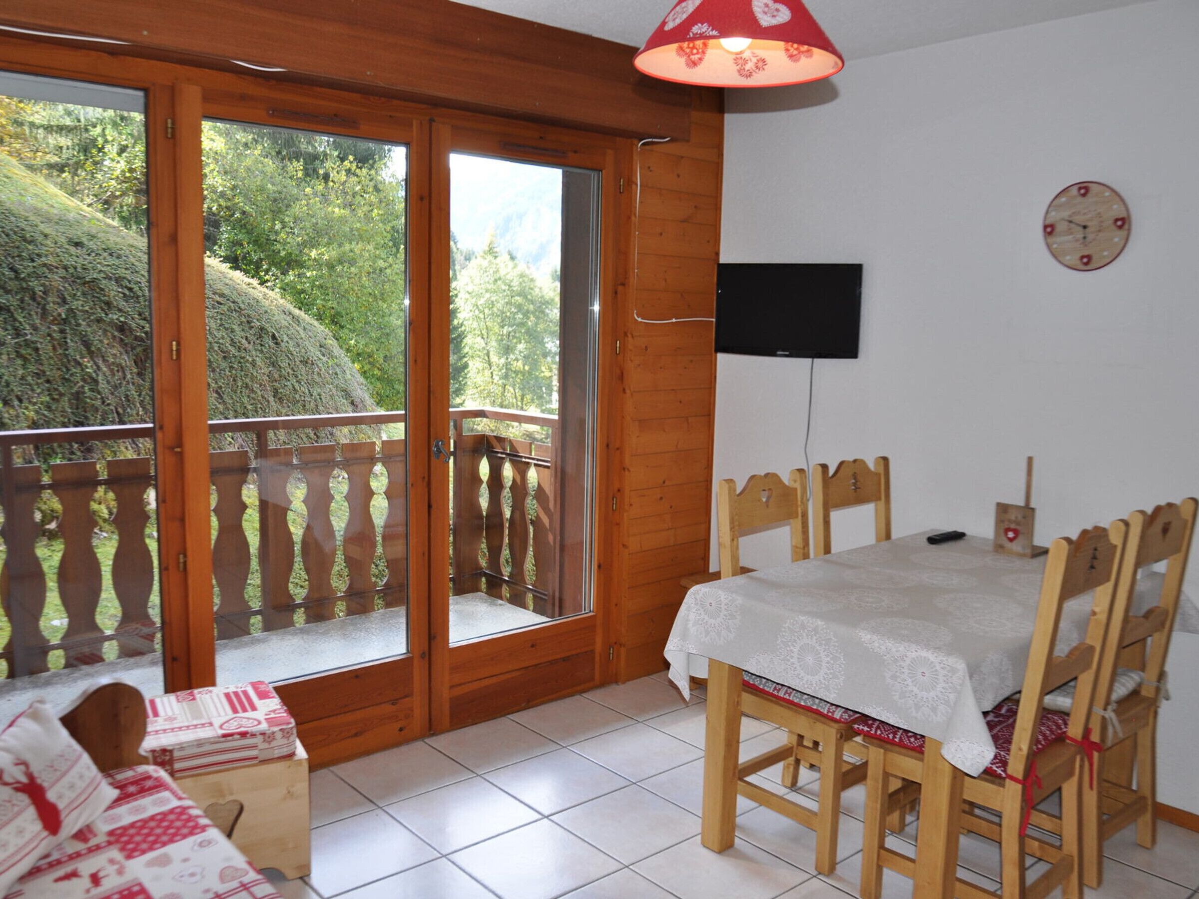 A room with a wooden balcony, a dining table with chairs, a TV mounted on the wall, and a bed with a patterned bedspread.