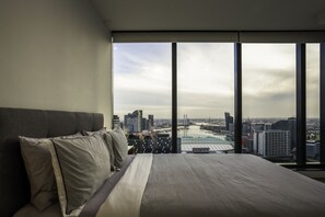 A modern bedroom with a large bed, a view of the cityscape through floor-to-ceiling windows, and a city skyline in the background.