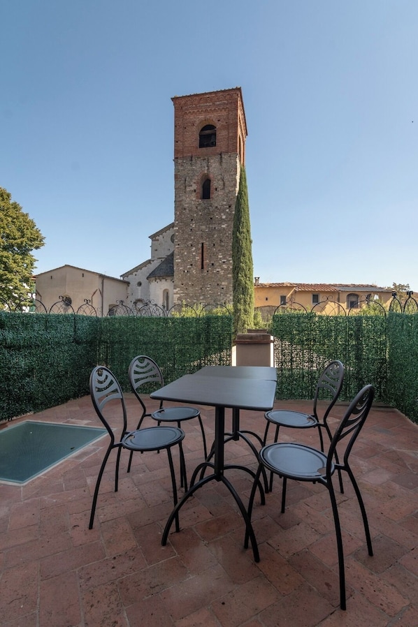 An outdoor patio with a table and chairs, a tall tower in the background, and a hedge in the foreground.