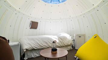 Um ambiente acolhedor com cama, uma mesinha, um pufe amarelo e um frigorífico compacto.