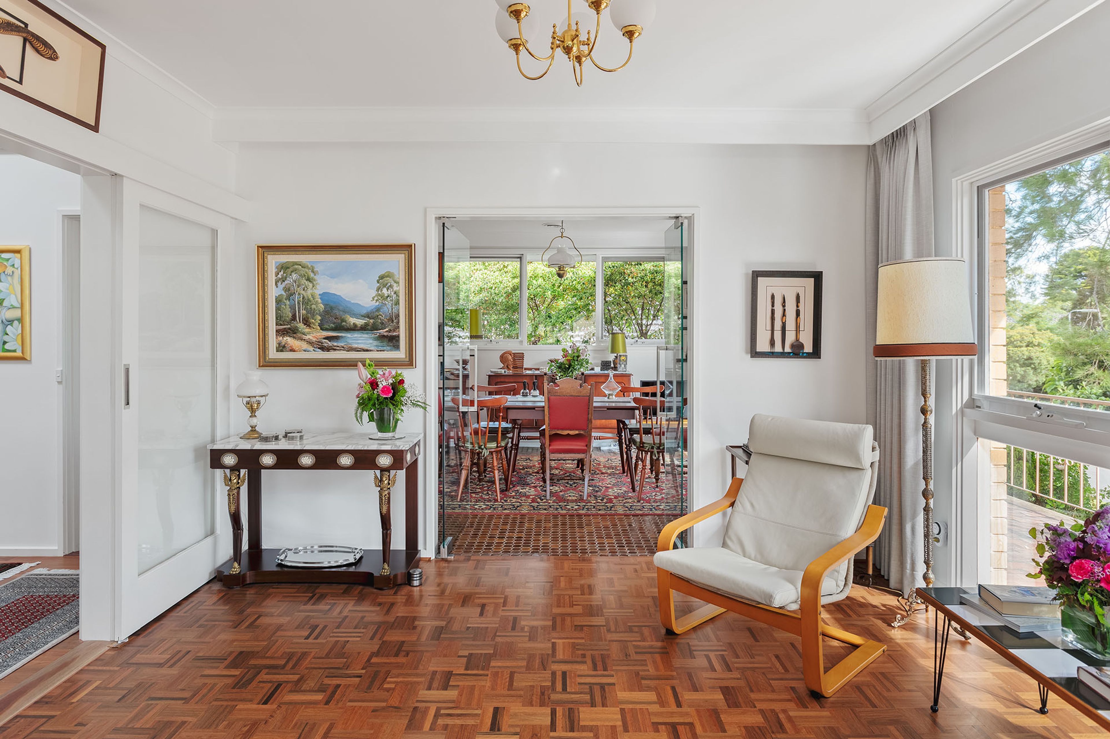A spacious room with a wooden parquet floor, a dining area with a red table and chairs, a white armchair, and a chandelier.