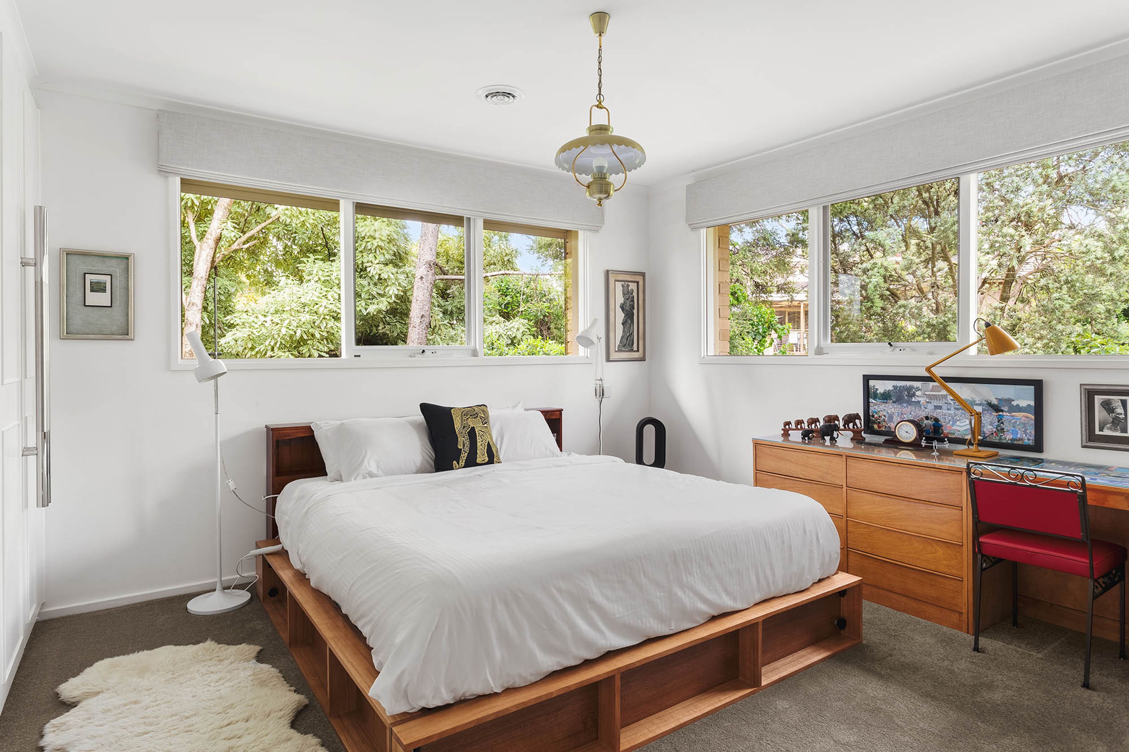 A bedroom with a bed, a desk, and a chair. There are windows and a view of trees.