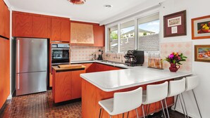 A modern kitchen with wooden cabinets, a stainless steel refrigerator, an oven, and a coffee machine. There is a window providing a view of the outside. - That 70s House in Mount Waverley (Mount Waverley)