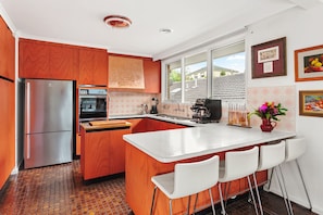 A modern kitchen with wooden cabinets, a stainless steel refrigerator, an oven, and a coffee machine. There is a window providing a view of the outside. - That 70s House in Mount Waverley (Mount Waverley)