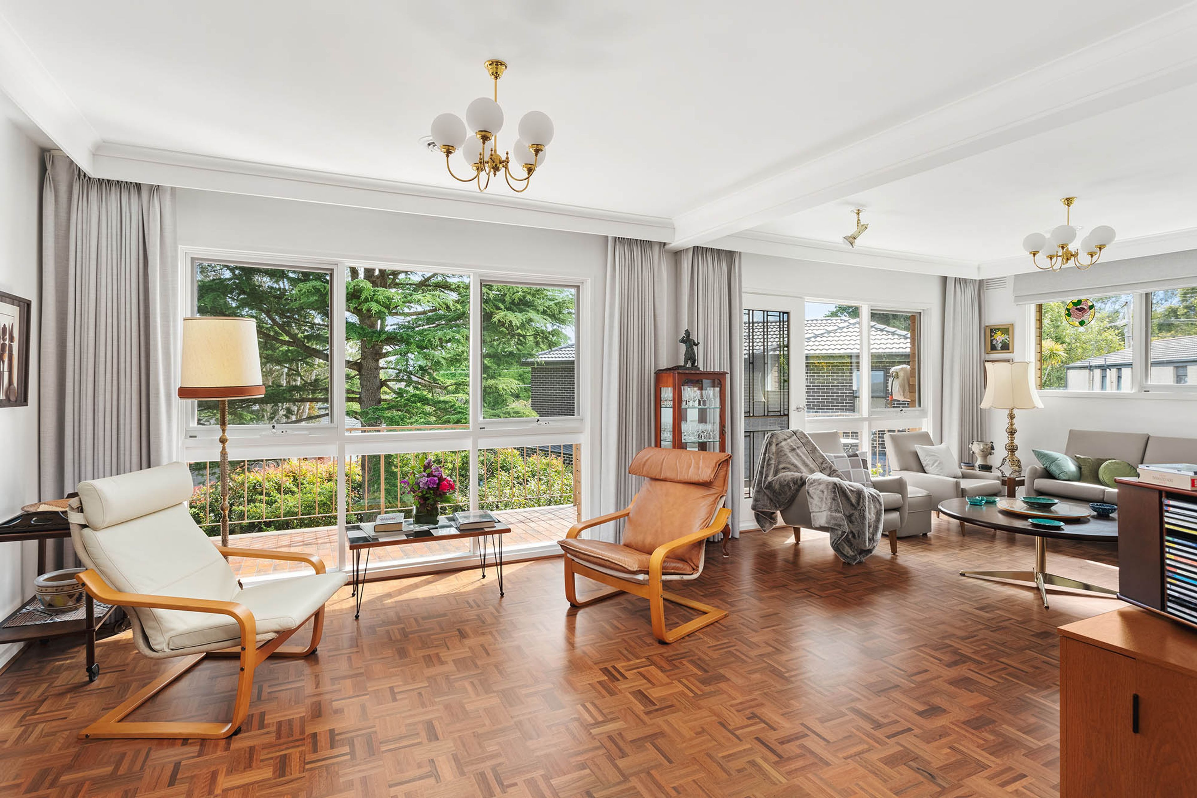 Amplia sala de estar con ventanas grandes, piso de madera en espiga y diversas opciones de asientos, incluyendo un sillón blanco y un sillón de cuero marrón.