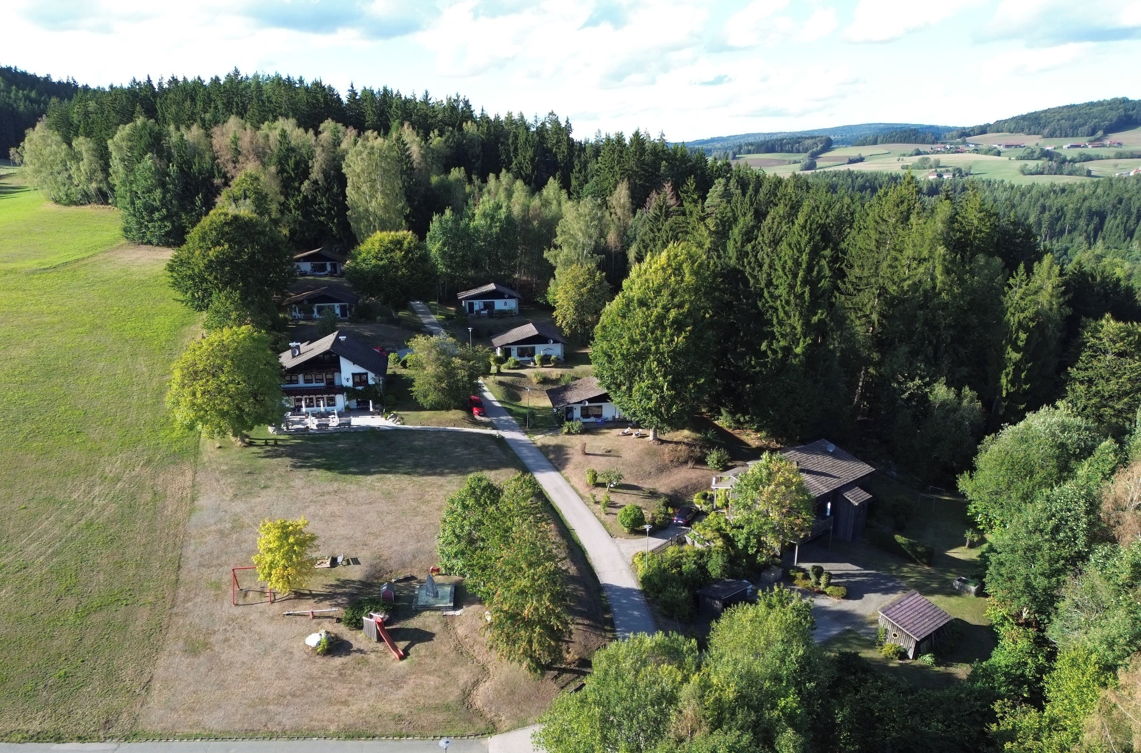 A rural property with multiple buildings, a paved driveway, and surrounding forested hills.