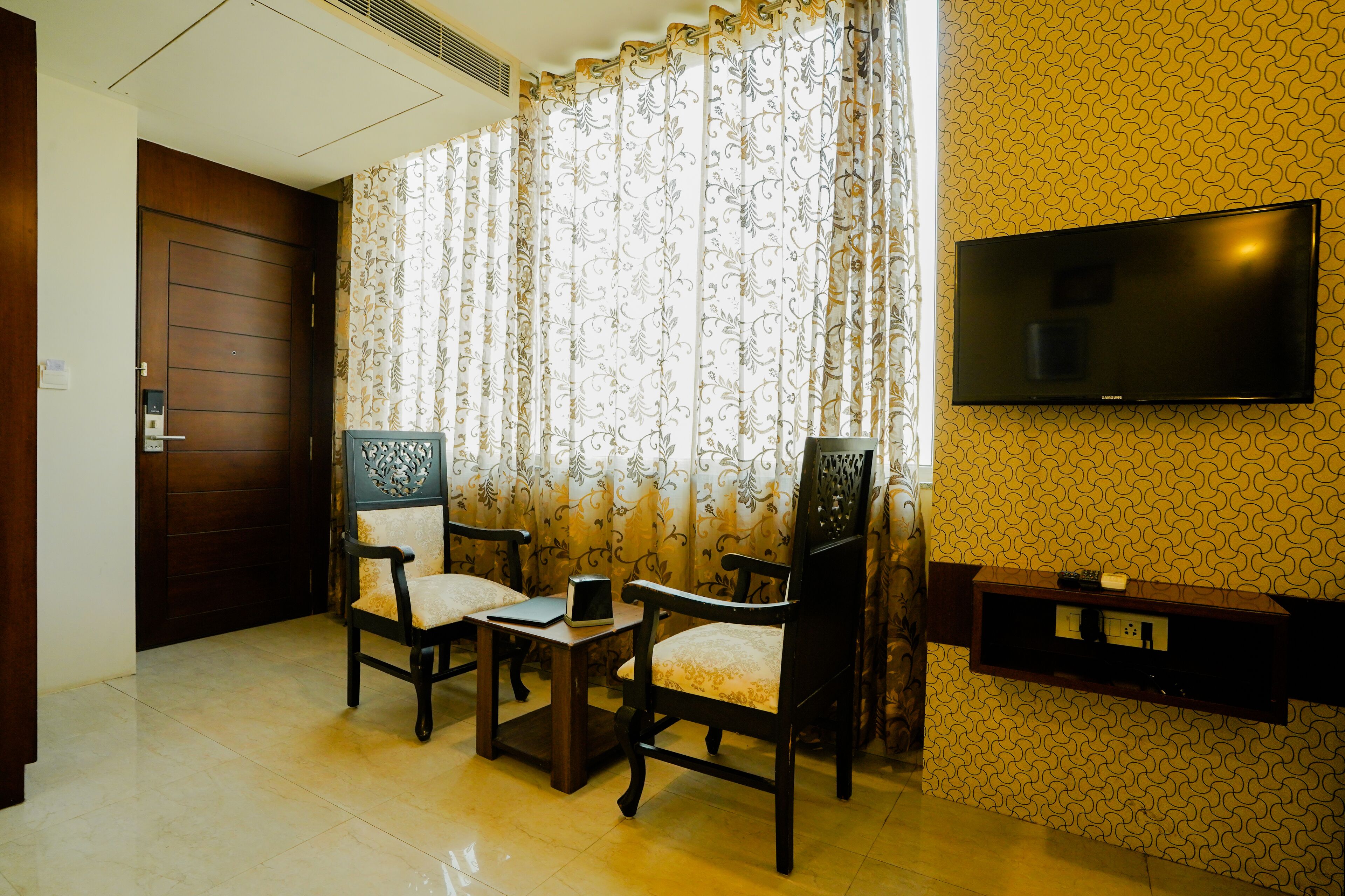 A hotel room with a wooden door, a TV mounted on the wall, two chairs, a small table, and a patterned wallpaper.