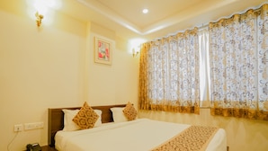 A hotel room with a large bed, a nightstand with a phone, a wall outlet, and a window with patterned curtains. - Hotel Cheelgadi (Jaipur)