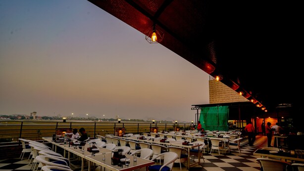 An outdoor dining area with tables and chairs, a view of the city, and a stage with a green backdrop. - Hotel Cheelgadi (Jaipur)