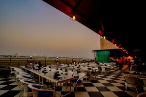 Un área de comedor al aire libre con mesas y sillas, vista a la ciudad y un escenario con fondo verde.