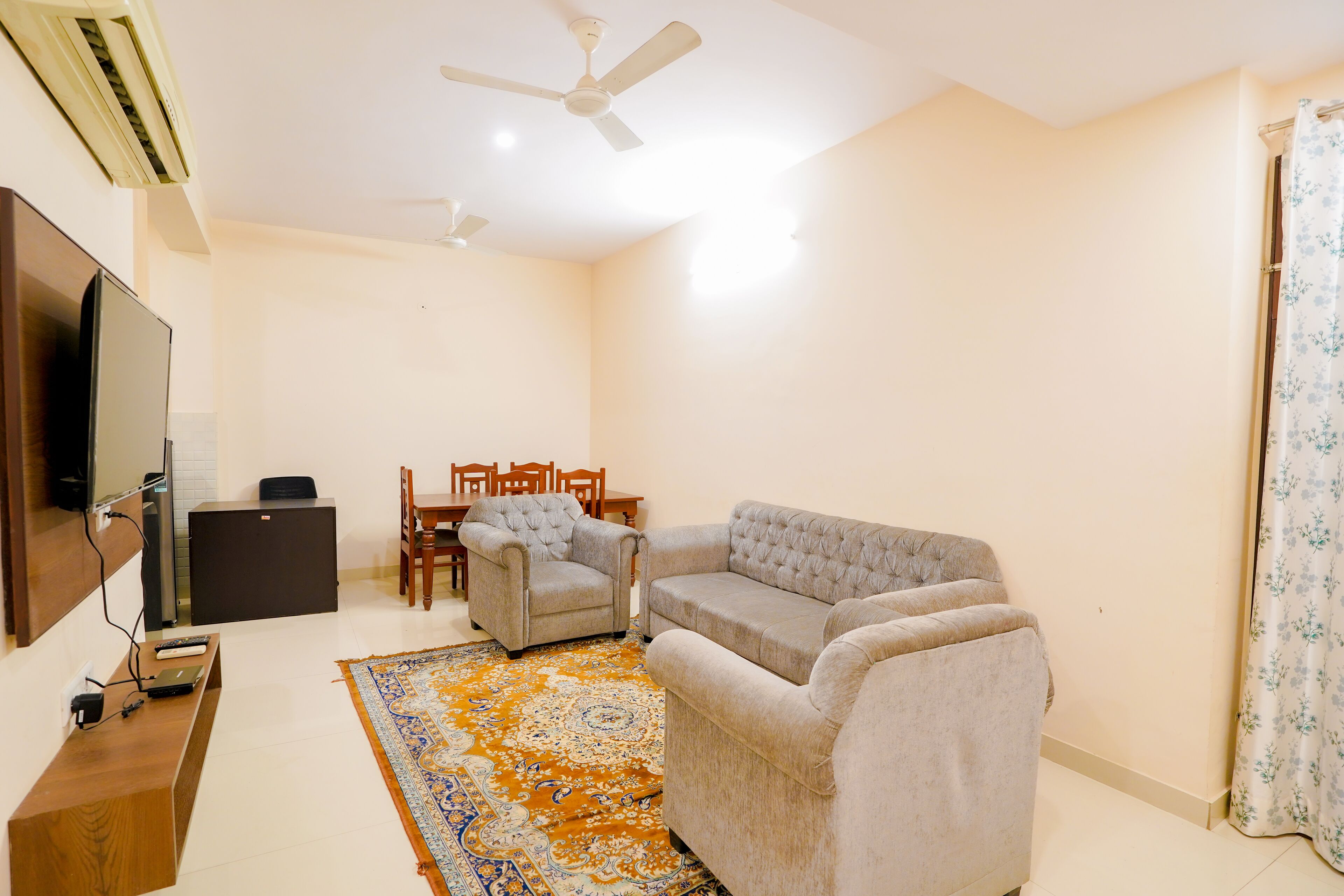 A living room with a sofa, armchairs, a TV, and a coffee table. There is a ceiling fan, a rug, and a window with curtains.