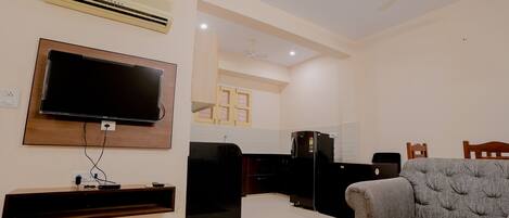 A living room with a flat-screen TV, a sofa, a coffee table, and a kitchen in the background.