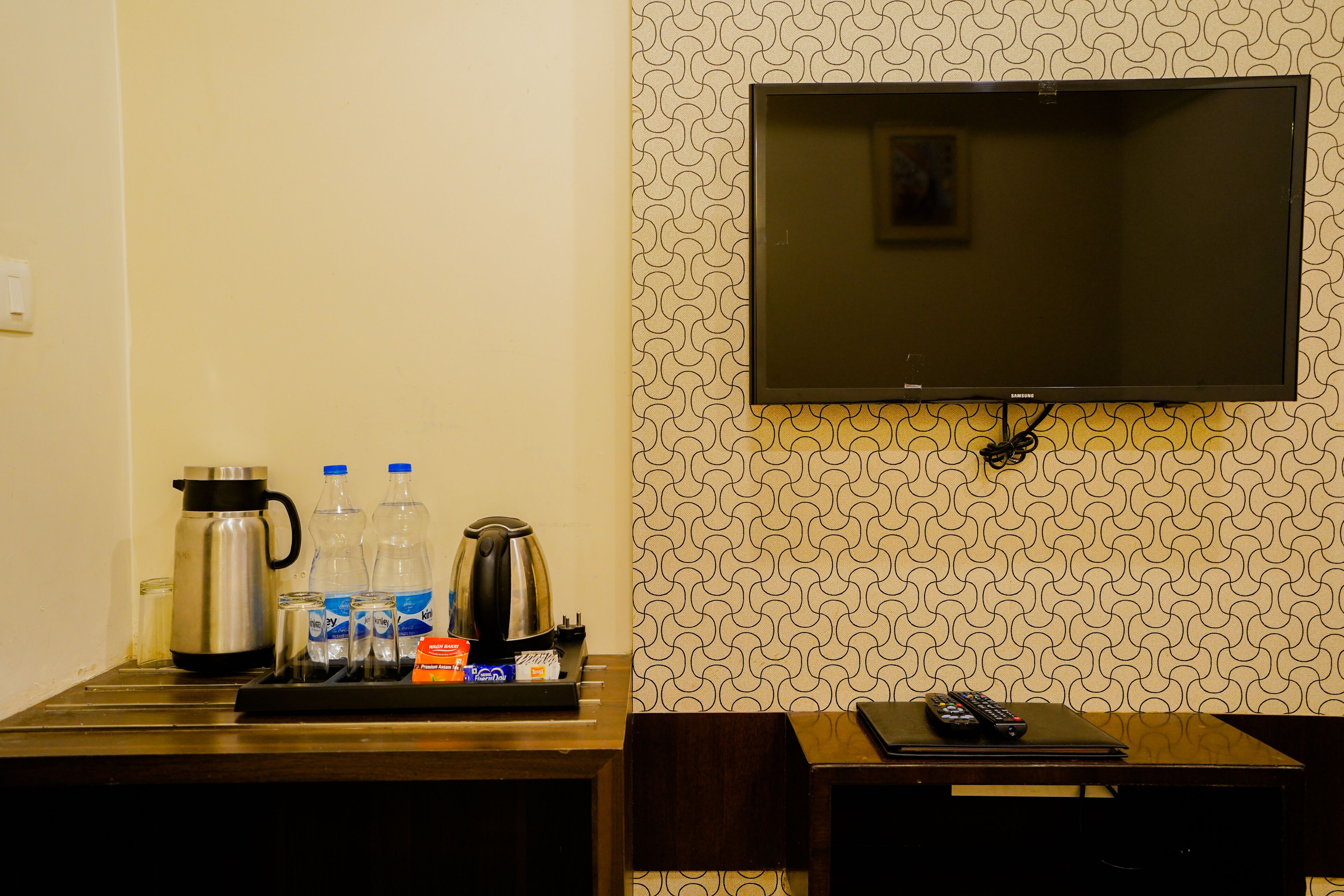 A hotel room with a TV mounted on the wall, a coffee maker, water bottles, and a tray with a kettle and cups.