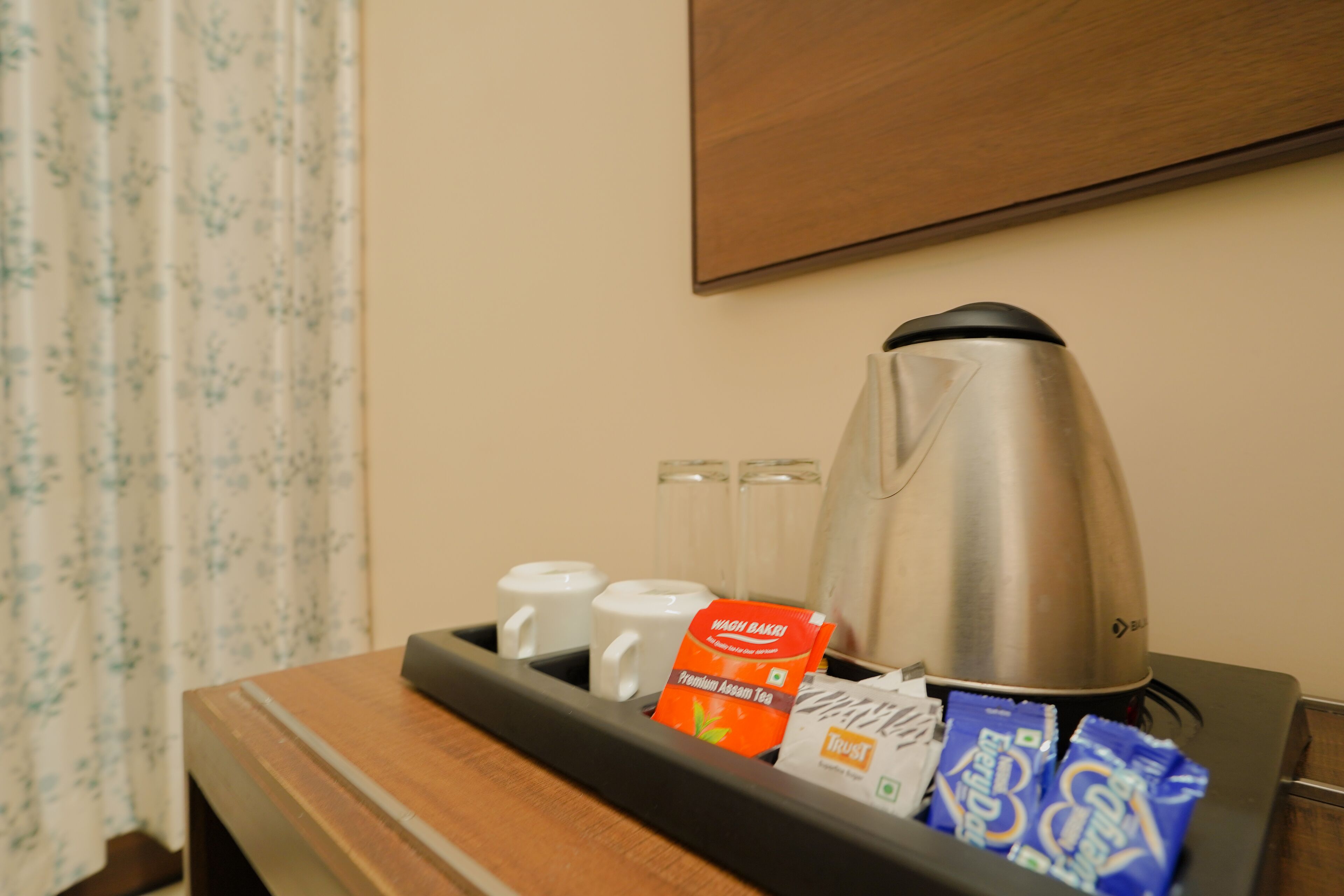 A tray with a stainless steel coffee pot, two white mugs, a packet of premium Assam tea, and several packets of instant coffee.