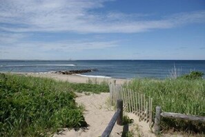 A sandy beach path leading to the ocean, with a wooden fence on the right and green vegetation on the left. - Town Neck Beach Retreat by the Marina (Sandwich)