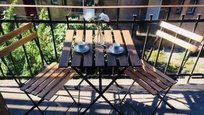 Un pequeño balcón al aire libre con una mesa de madera y dos sillas, un jarrón con flores blancas, y vistas a zonas verdes y edificios al fondo.