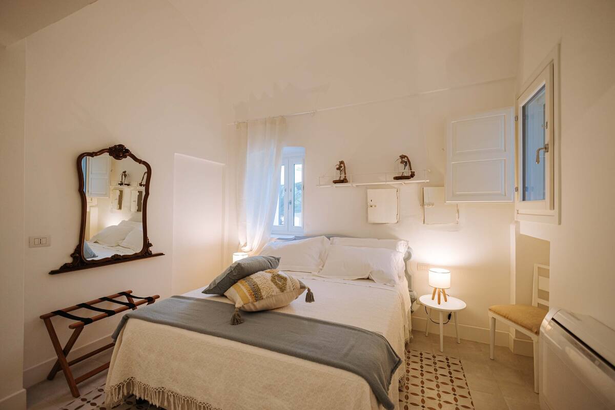 A bedroom with a bed, a bedside table with a lamp, a chair, a mirror, and a shelf with decorative items.