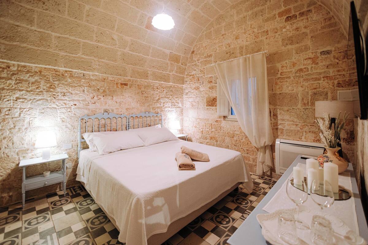 A bedroom with a bed, a nightstand, a bedside table, a mirror, and a window with curtains.