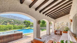 Un espacio exterior techado con un jacuzzi, asientos y vistas a una piscina y montañas.