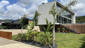 Una casa moderna de dos pisos con techo plano, ventanas amplias y un jardín cuidado con diversas plantas y una cerca de madera.
