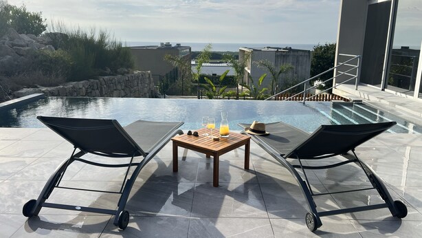 Un área junto a la piscina con dos sillones de descanso, una mesa con bebidas y vistas al océano y edificios.