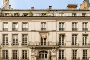 Ein mehrgeschossiges Gebäude mit einer symmetrischen Fassade, das gleichmäßig verteilte Fenster, dekorative Eisenverzierungen an Balkonen und einen zentralen Eingang mit Bogen aufweist.