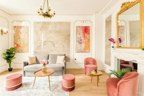 Una sala de estar moderna con un sofá gris, sillones rosas y una chimenea. Hay cuadros abstractos en las paredes y un candelabro sobre el sofá.