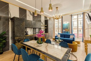 A modern living room with a blue sofa, a dining table with chairs, and a chandelier.