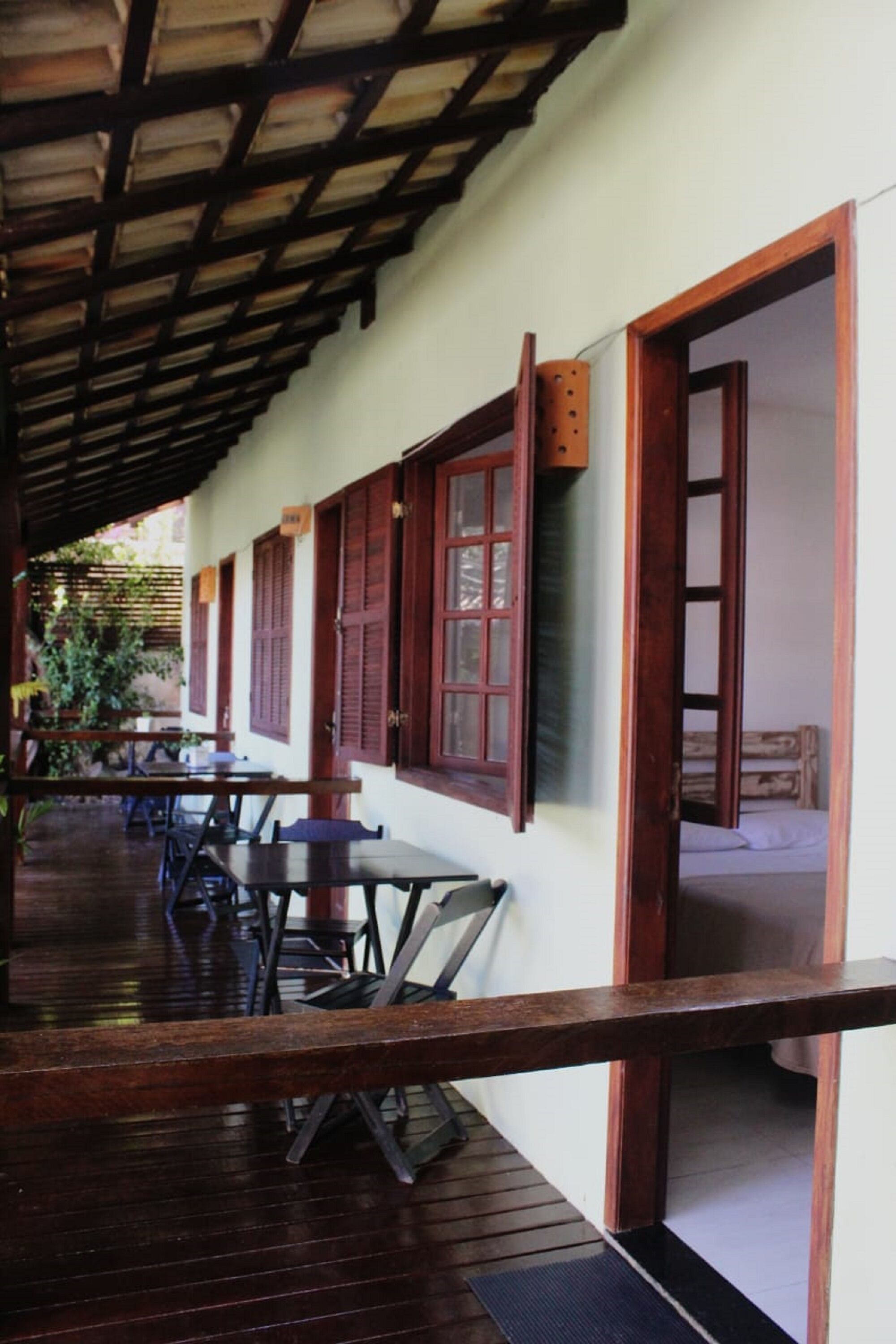 A covered outdoor seating area with wooden tables and chairs, a wooden railing, and a view of a room with a bed.