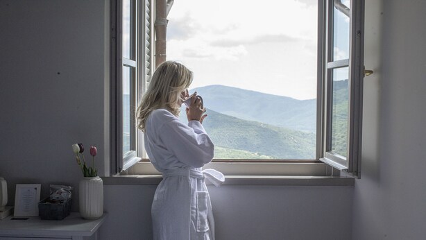 Una persona vestida de blanco de pie junto a una ventana con vistas a montañas.