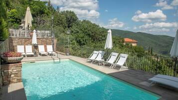 Une piscine entourée de chaises longues et de parasols, bordée de murs en pierre et de verdure, avec une vue sur des collines en arrière-plan.