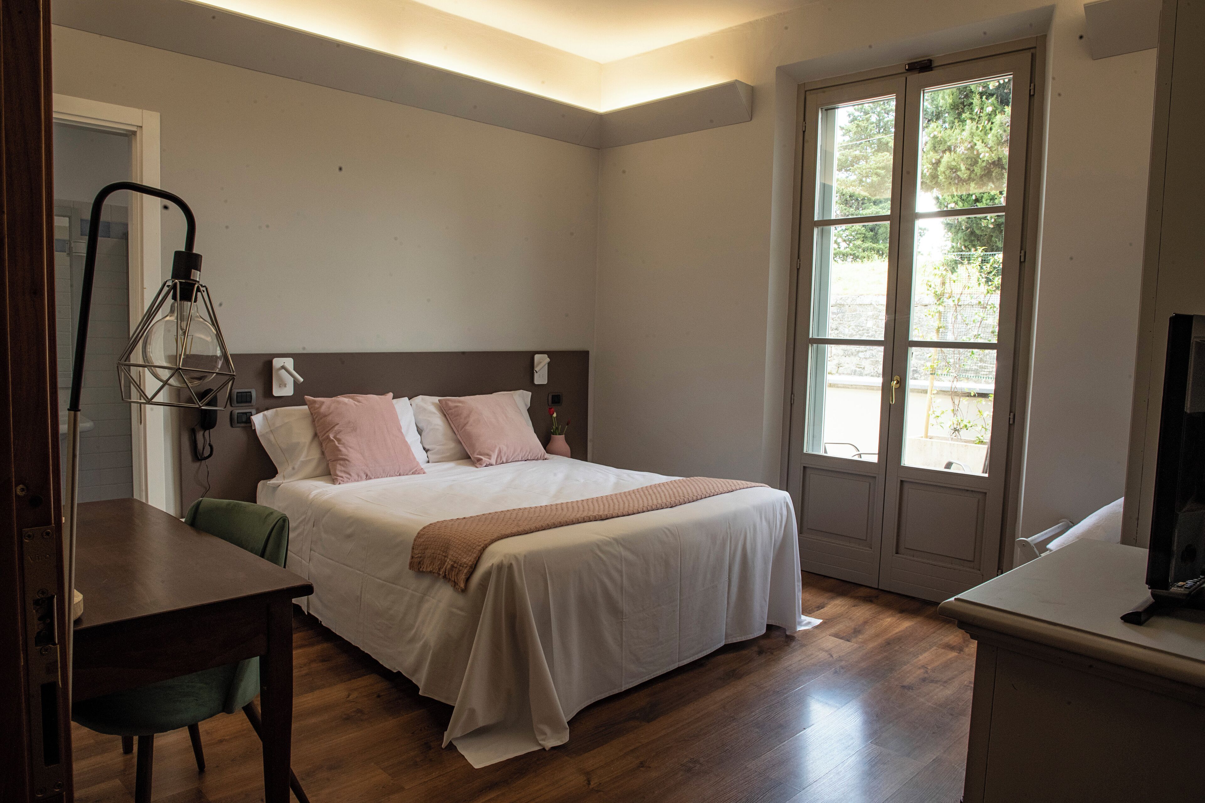 Quarto com cama, mesa de cabeceira, cadeira e janela com vista para vegetação.