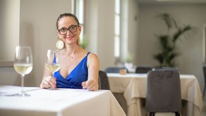 Una persona sentada en una mesa con una copa de vino blanco, en una habitación con mesas y sillas, y una planta al fondo.