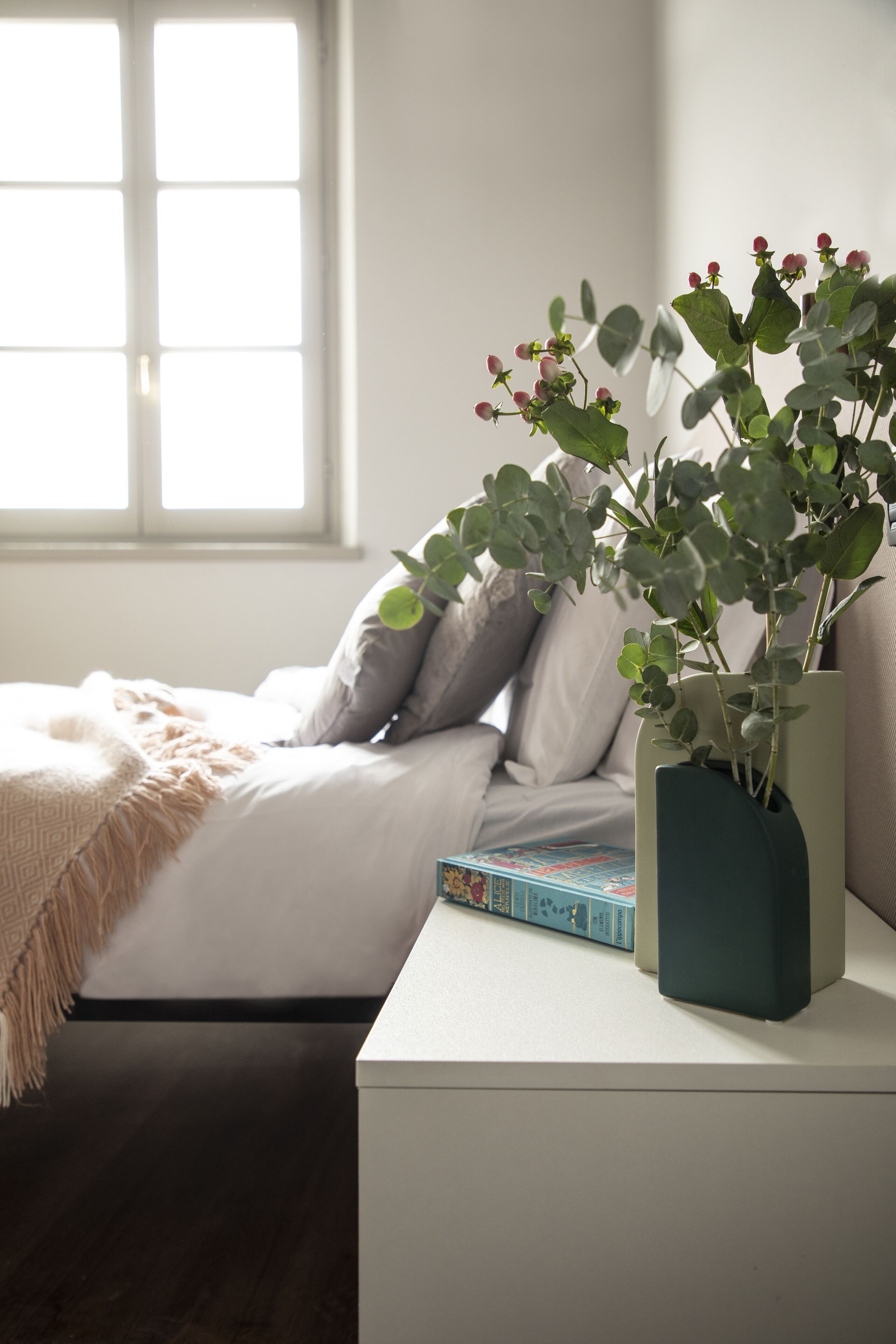 A neatly made bed with a white duvet, a bedside table with a book, and a window with natural light.