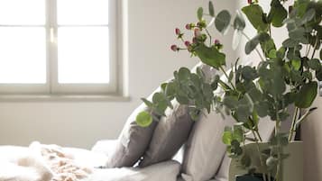 Una cama bien hecha con edredón blanco, una mesita de noche con un libro y una ventana con luz natural.