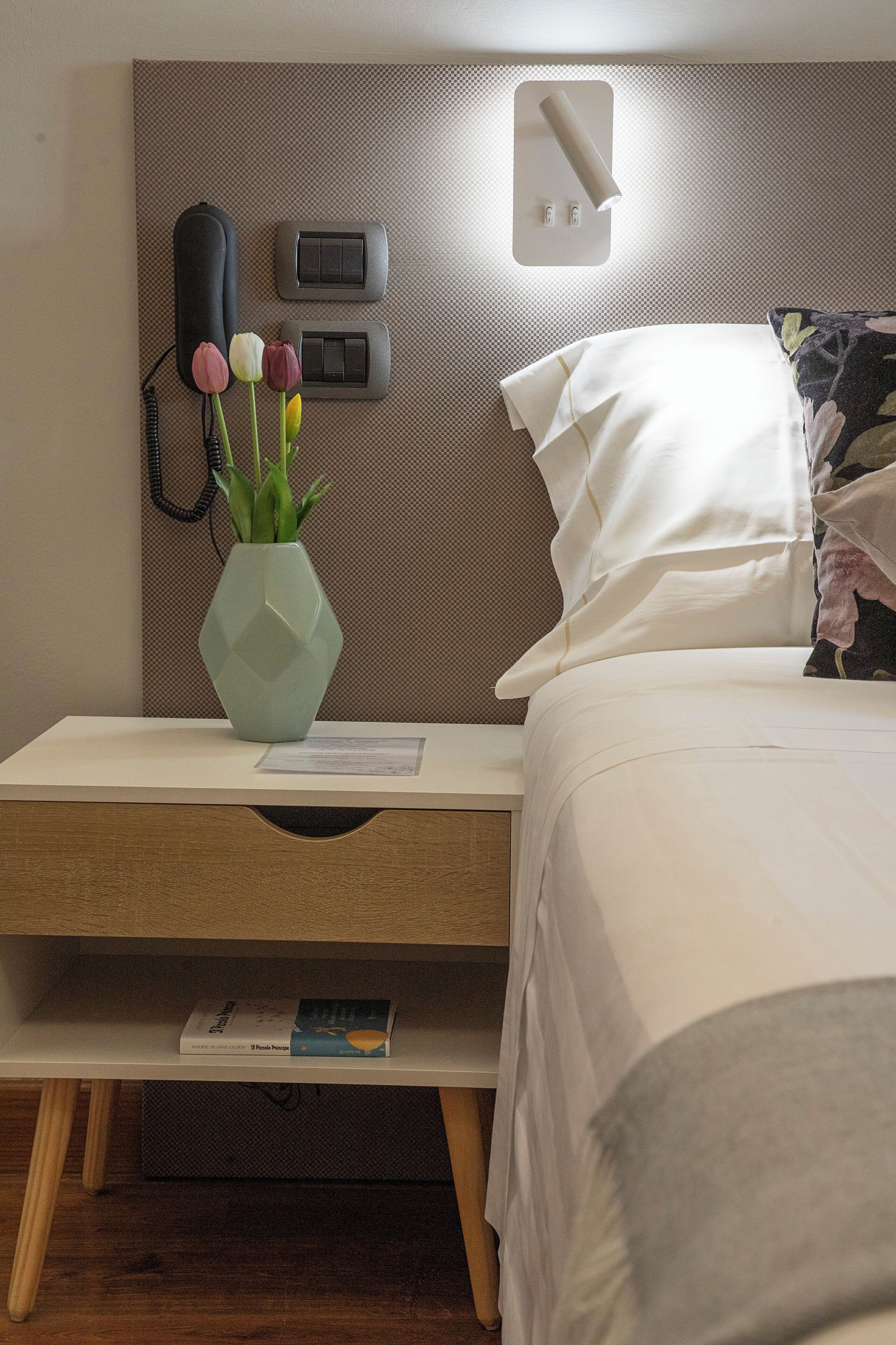 A neatly arranged hotel room with a bed, a bedside table with a vase of flowers, a lamp, and a wall-mounted phone.