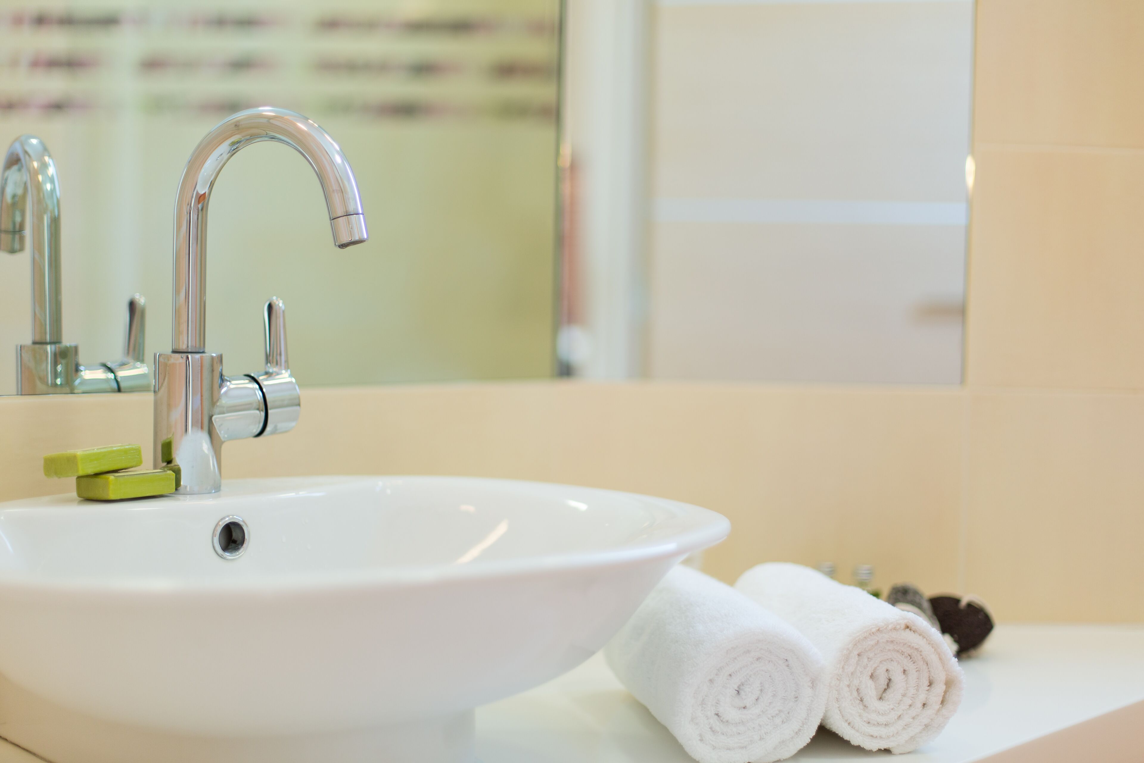 Ein weißes Waschbecken mit chromfarbenem Wasserhahn und einem grünen Seifenspender. Auf der Ablage liegen zwei gerollte weiße Handtücher.