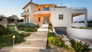 A modern two-story house with a stone pathway leading to the entrance, surrounded by lush greenery and a well-maintained garden. - Deluxe Villa Ianthos - Outdoor Hot Tub & Kids Pool (Rethymno)
