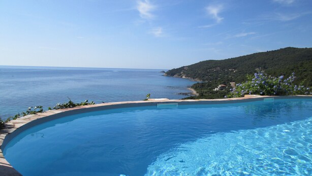 Une piscine d’un bleu éclatant, à bords incurvés, entourée d’une terrasse en pierre et offrant une vue sur la mer et les collines.