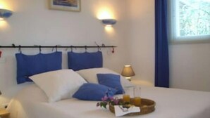 Une chambre à coucher comprenant un lit, des oreillers, une table de chevet, une lampe, une fenêtre avec des rideaux et une œuvre d’art accrochée au mur.