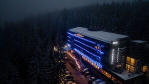 A multi-story hotel with a modern design, featuring balconies with blue lights, a snow-covered roof, and a parking area in front. - Hotel Cindrel (Sibiu)
