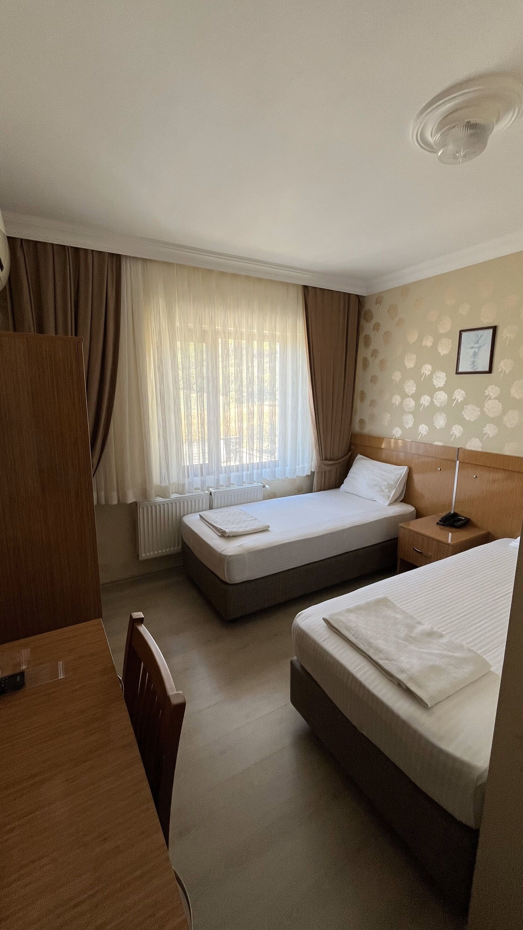 Habitación de hotel con dos camas, un escritorio, una silla, un radiador y una ventana con cortinas.