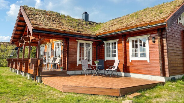 Eine Holzhütte mit geneigtem Dach, einer Terrasse mit Sitzgelegenheit und Blick auf einen grasbewachsenen Hügel.
