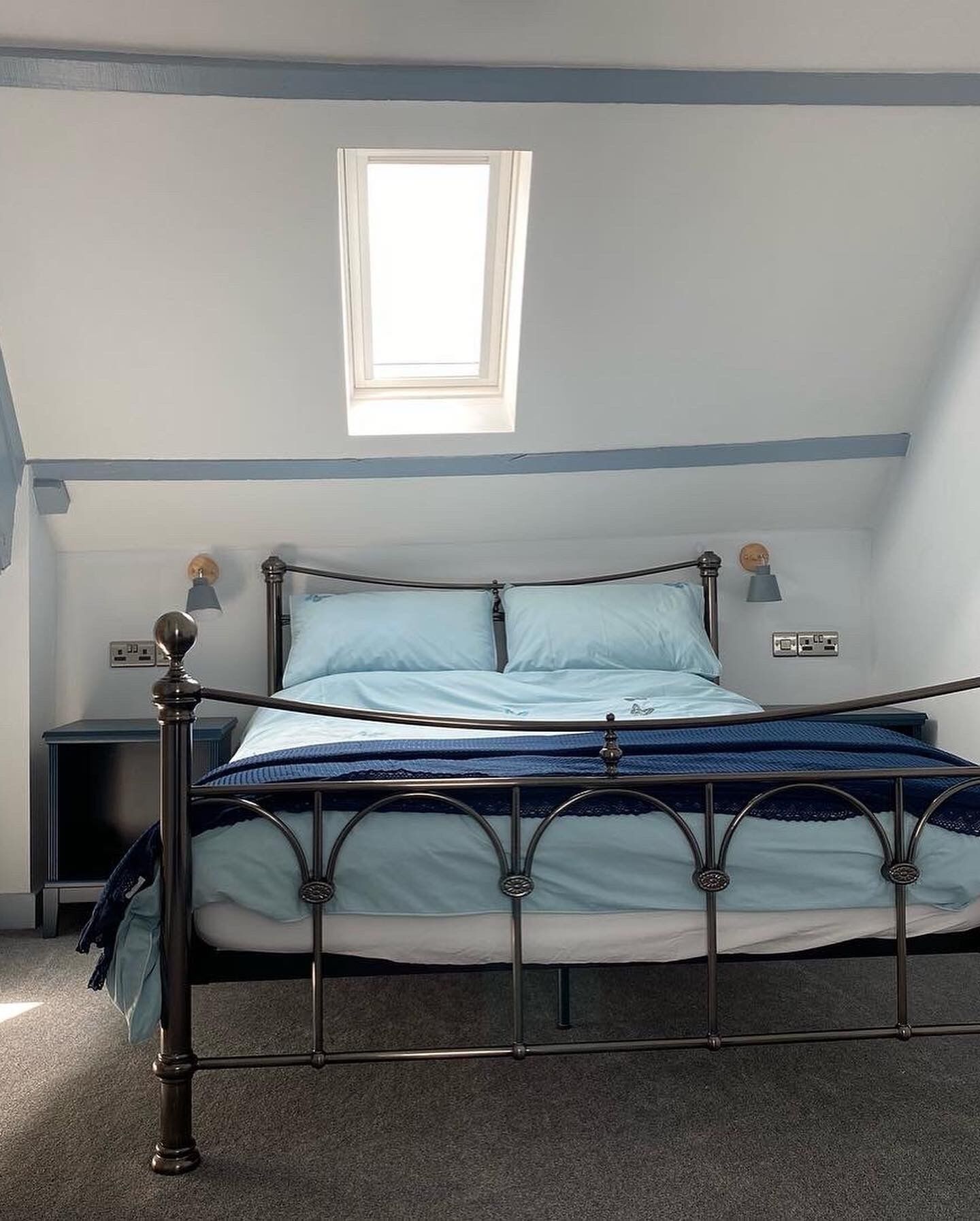 A bedroom with a metal bed frame, a small bedside table, a wall socket, and a window with natural light.
