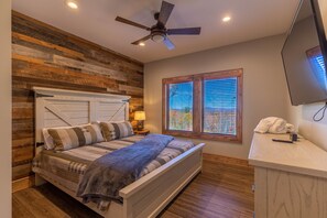 A bedroom with a wooden headboard, a bed with striped bedding, a ceiling fan, a nightstand with a lamp, and a window with blinds.