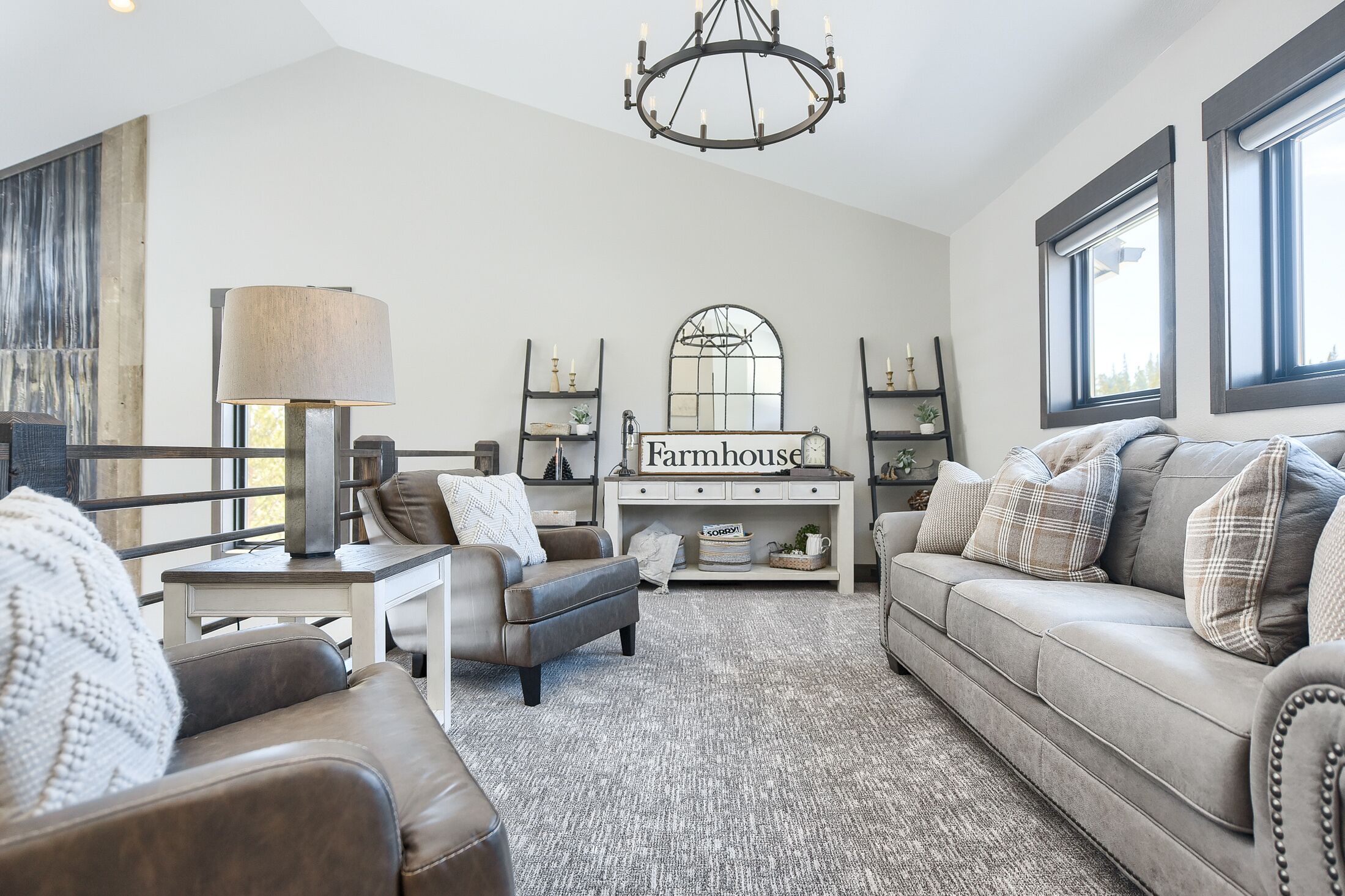 A spacious living room with a grey sofa, a brown leather armchair, and a white table with a lamp. A chandelier hangs from the ceiling, and there are two windows with blinds.
