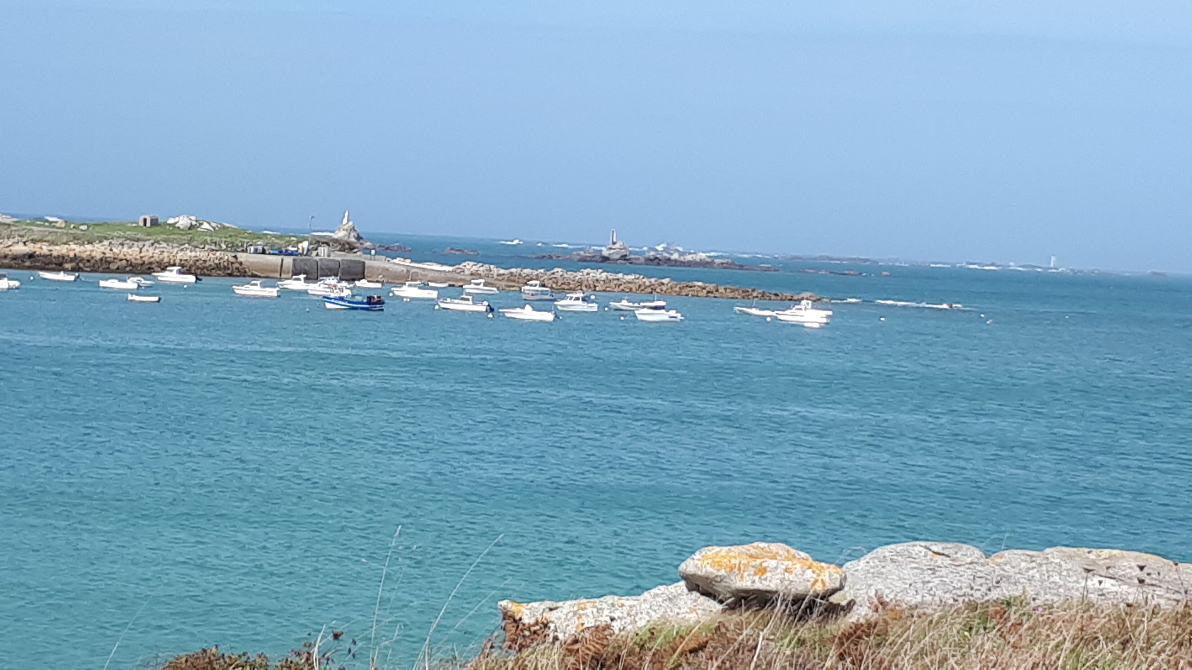 Paisaje costero con barcos amarrados cerca de una costa rocosa y un faro al fondo.